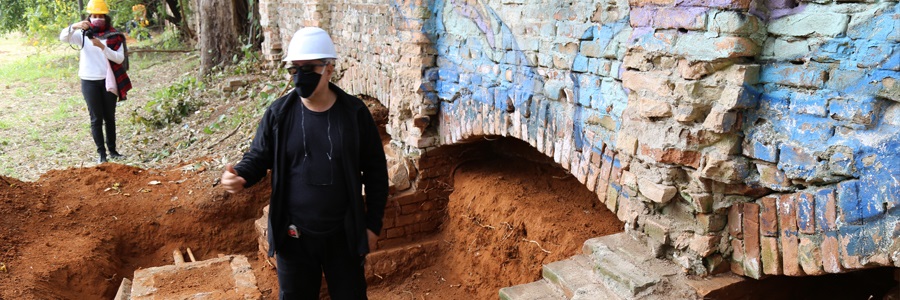 Foto de uma escavação de um muro com terra vermelha com dois trabalhadores de capacete 